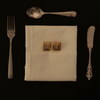 Set of silverware including a knife, spoon, and fork on a white napkin with gold embroidery against a black background.